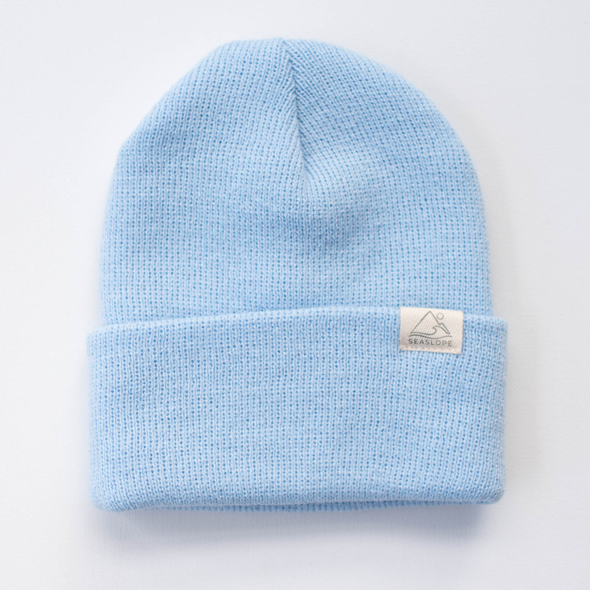 This light blue infant/toddler beanie features a folded brim and a subtle mountain design tag. Crafted from 100% acrylic, it offers a soft and stretchy fit. The beanie is displayed on a plain white background.