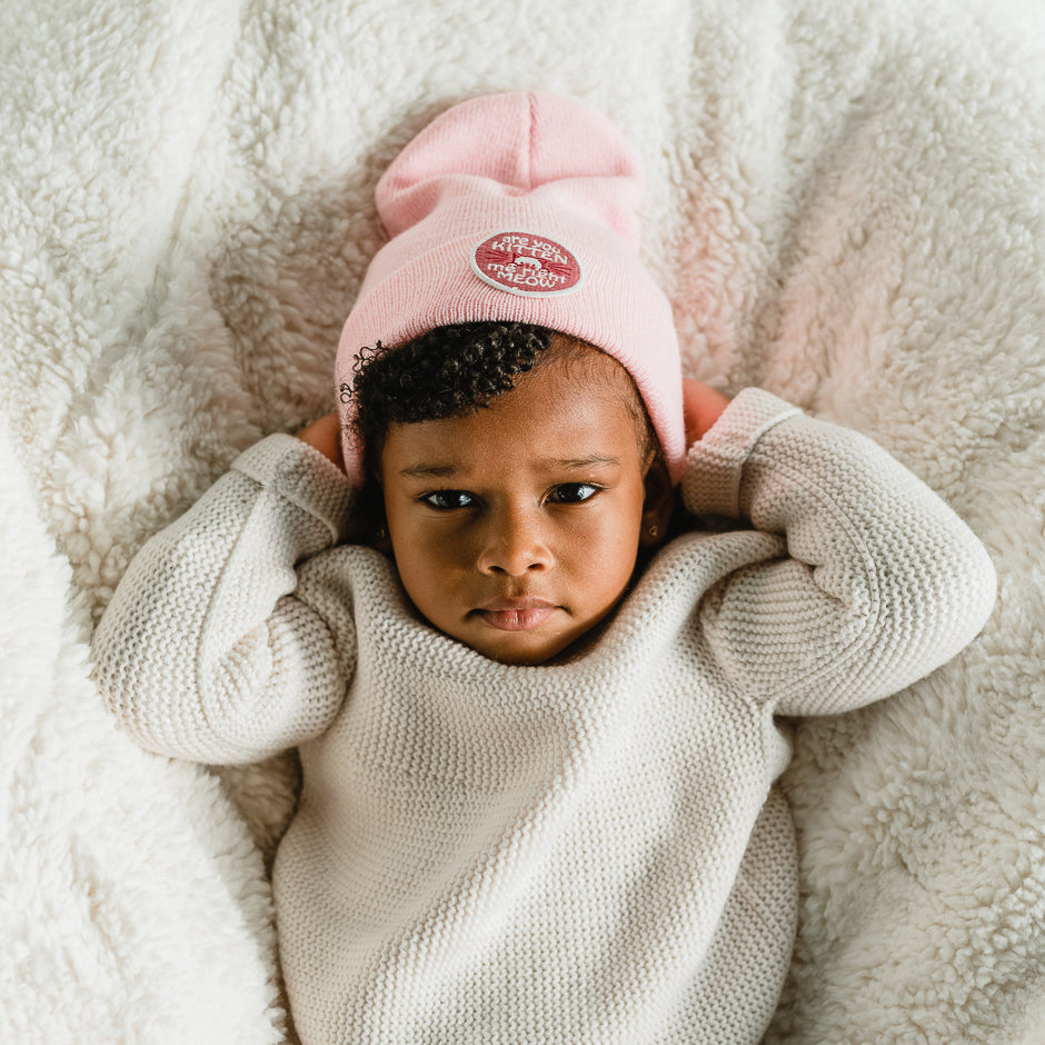 A young child in a cozy cream sweater and a playful beanie lies on a soft, fluffy surface. With arms raised above the head, the child gazes calmly at the camera.