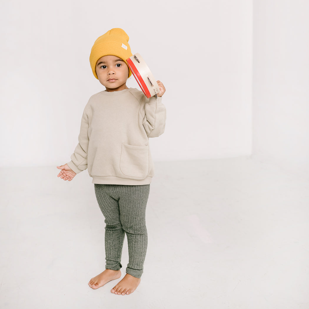 A young child is wearing a snug beanie along with a beige sweatshirt and green pants, holding a tambourine while standing barefoot on a light-colored floor. The beanie offers soft stretch comfort as it is made from 100% acrylic. The scene is set against a plain white backdrop, adding to the minimalist aesthetic.