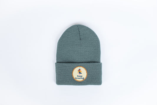 A spruce-colored kids' beanie featuring a folded brim and a circular patch with a stylized plant design and the words “Keep Growing” is centered against a white background.