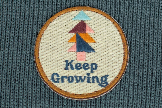 The Spruce Kids Beanie is adorned with an embroidered patch that features an abstract tree design in pastel colors. The green fabric provides a vibrant backdrop, enhancing the design's visual appeal. Below the tree, the motivational message "Keep Growing" is stitched, complementing the overall theme of growth and positivity.