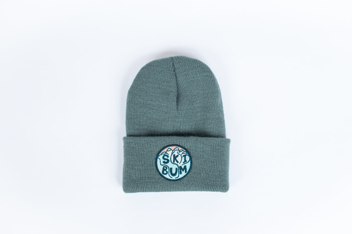 The Ski Bum - Spruce Kids Beanie is a stylish gray knit beanie featuring a folded brim, perfect for infants or toddlers. It includes a circular patch with "SKI BUM" in blue letters, which stands out against the white background that highlights its texture and color.