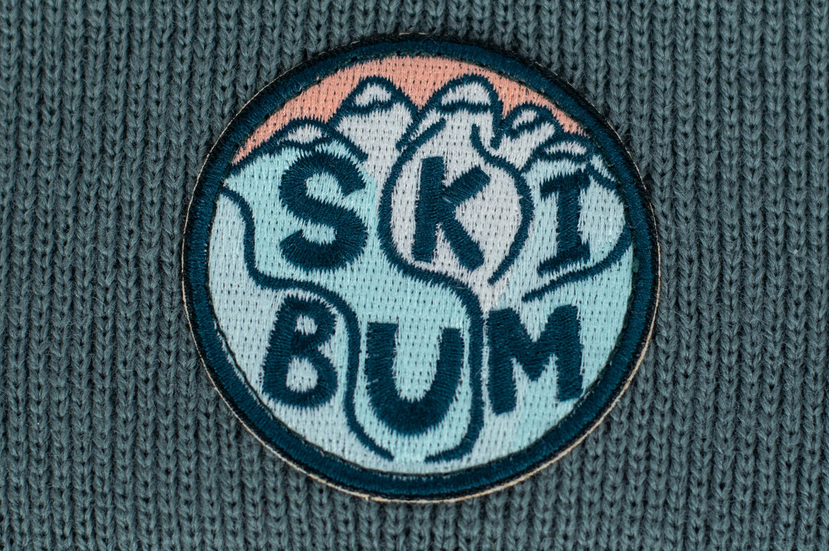 The Spruce Kids Beanie by seaslope1 is a delightful accessory for toddlers, highlighting a circular knitted patch with the bold statement "SKI BUM." Its cozy knit design is enhanced with abstract mountain shapes in teal, pink, and white on textured fabric, adding an extra layer of charm.