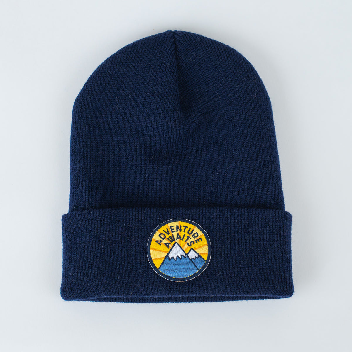 The Adventure Awaits Navy Beanie, available for both youth and adults, showcases a folded brim and an embroidered patch with blue mountains beneath a yellow sky, accompanied by bold "Adventure Awaits" lettering. Perfect for style-conscious individuals, this beanie seamlessly combines fashion with functionality.