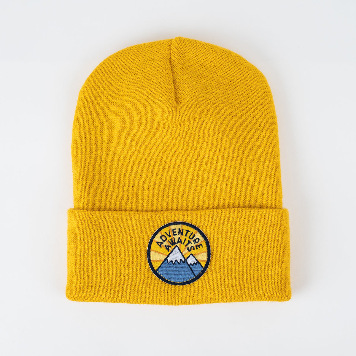 The Adventure Awaits beanie is a stylish yellow knit hat with a folded brim. It features an embroidered circular patch with mountains and the phrase "Adventure Awaits" on the front. This handcrafted beanie is displayed against a simple white backdrop.