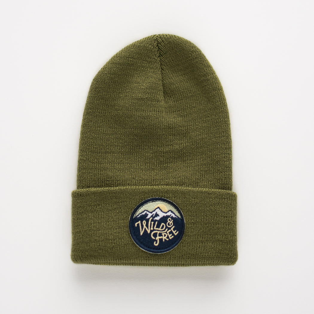 Introducing the Wild & Free - Moss Kids Beanie: an olive green toddler accessory with a folded cuff that features a round patch. The patch displays a mountain design, highlighting "Wild & Free" in white embroidery, making this charming piece stand out against a plain white background.