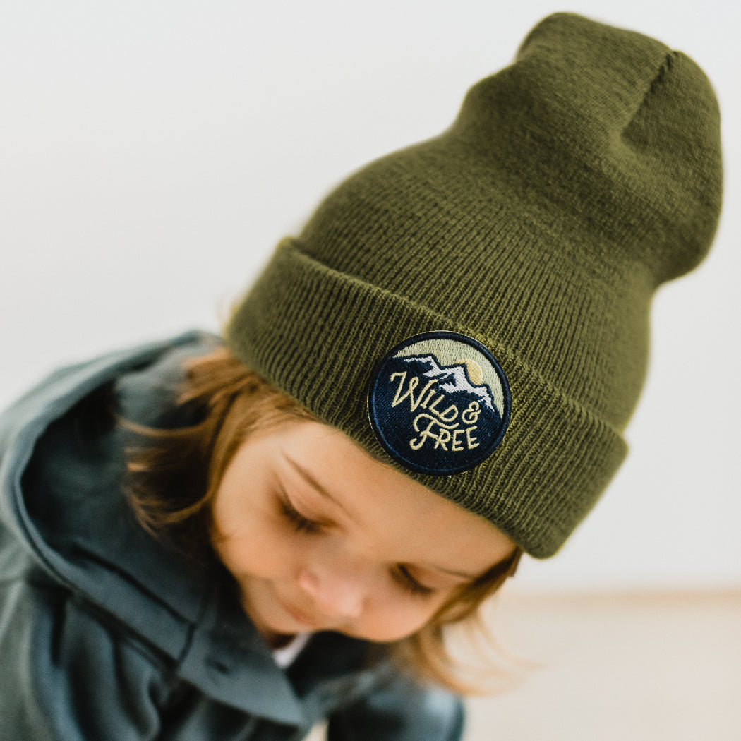 A young toddler is wearing a moss-colored beanie with a "Wild & Free" patch and a mountain design. The child is looking downward, cozy in a grey-green jacket.