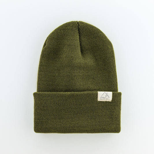 This olive green knit beanie features a folded brim and is adorned with a small rectangular white patch on the front. Enjoy its soft, stretchy fit, crafted with care in the USA. The beanie is displayed on a light gray background.