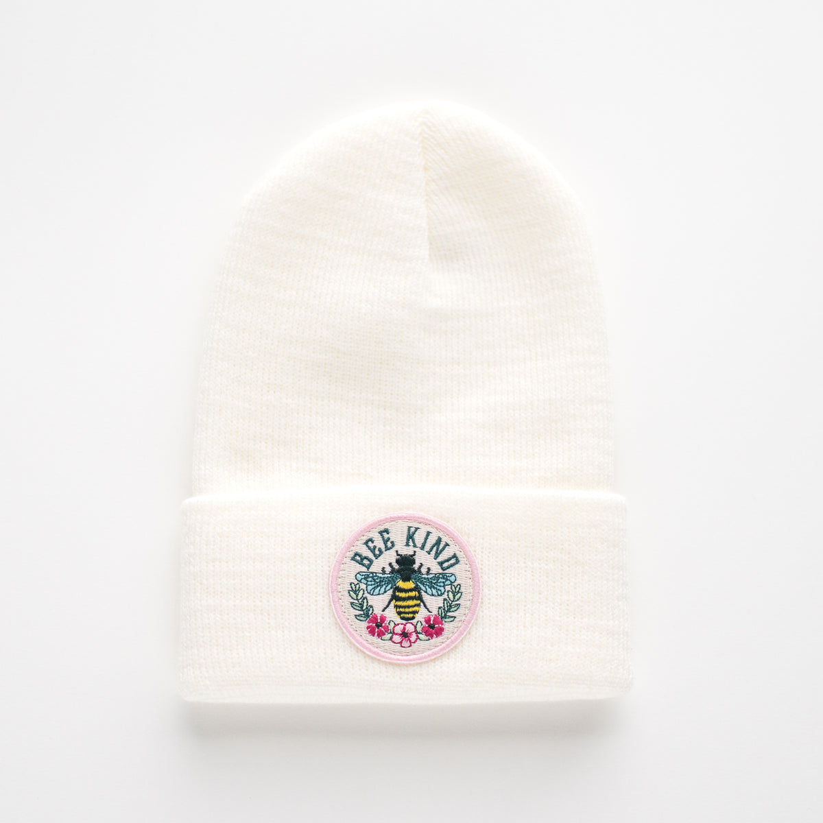 The "Bee Kind - White Kids Beanie" is a USA-made accessory featuring 100% acrylic material. It includes a folded brim and displays an embroidered circular patch with a bee illustration surrounded by pink flowers and the message "Bee Kind.