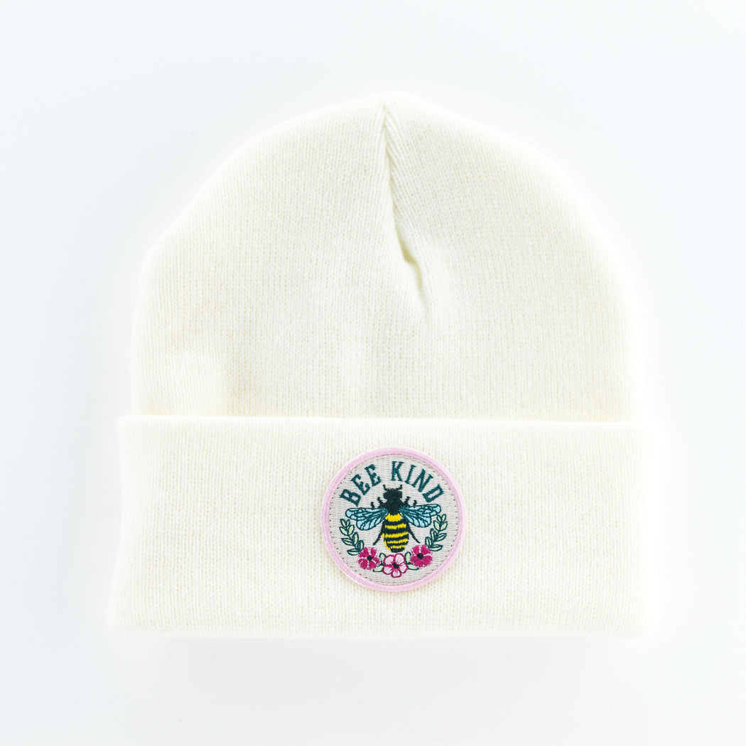 This cream-colored knit beanie is designed for adults and features a front patch adorned with a bee surrounded by flowers. The words "Bee Kind" are prominently displayed, offering warmth and style.