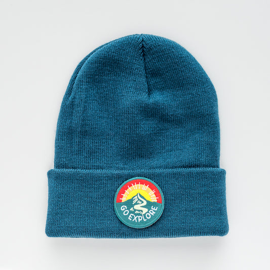 A stylish teal knit beanie, handmade with care, is displayed on a white background. This "Go Explore | Glacier - Adult Beanie" features a round patch adorned with the words "GO EXPLORE" and an image of a person hiking against a multicolored sunset.