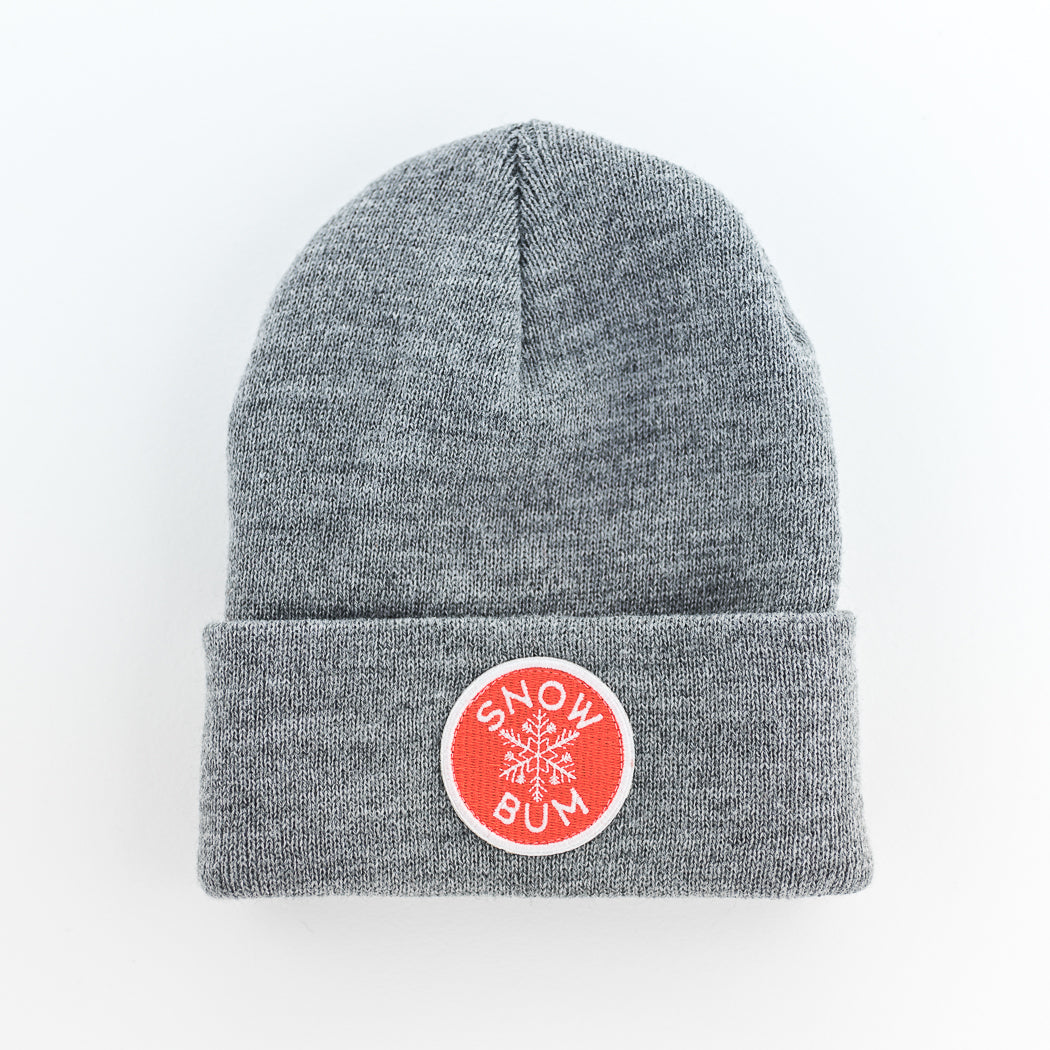 The handmade knit beanie in gray features a folded brim and a red circular patch with a white snowflake design and the text "SNOW BUM" on the front.