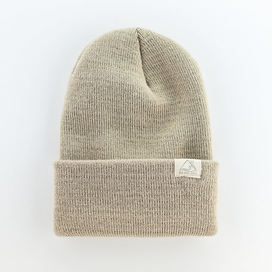 The SAND INFANT/TODDLER BEANIE is a soft, stretch-fit knit beanie in beige. It has a folded brim and is made in the USA. The beanie is showcased against a plain white background.