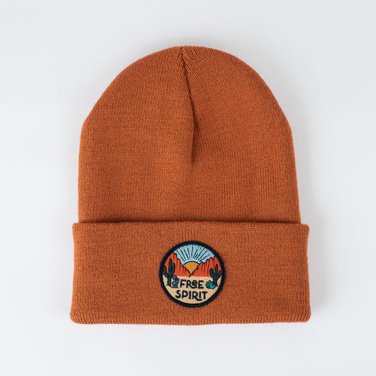 The "Free Spirit | Canyon - Adult Beanie" is an orange knit beanie with a folded brim. It includes a circular patch showing a sunrise over mountains, framed by cacti, and the words "FREE SPIRIT" at the bottom. This insulated beanie is ideal for adventurers and is presented against a plain white background.