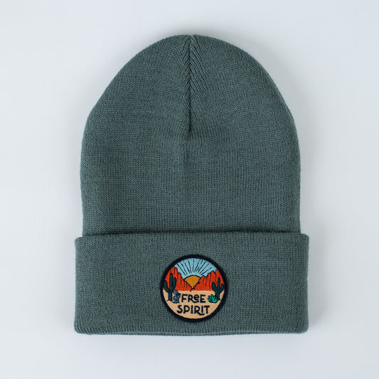 This insulated seafoam beanie features a vibrant patch on the front fold that depicts a desert scene with cacti, mountains, a sunburst, and the phrase "Free Spirit." Its knit texture offers warmth and style, creating a striking contrast against a plain white background.