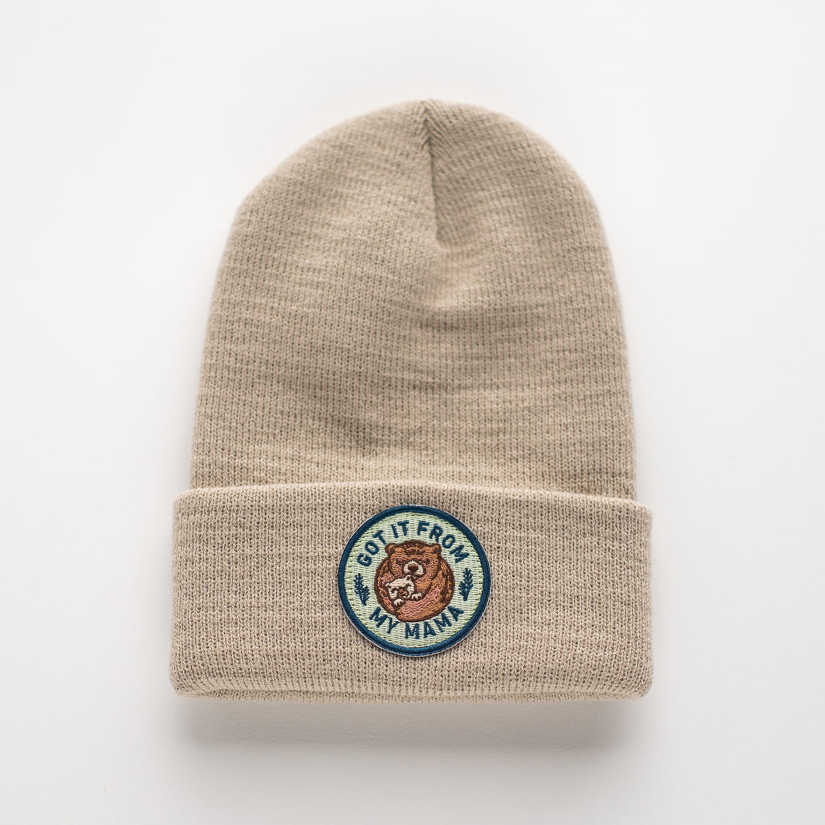 The Sand Kids Beanie by seaslope1 is a beige knit hat with a fold-over cuff. It features an embroidered patch on the front, showcasing a bear's face with the phrase "Got It From My Mama." It's proudly made in the USA for quality and style.