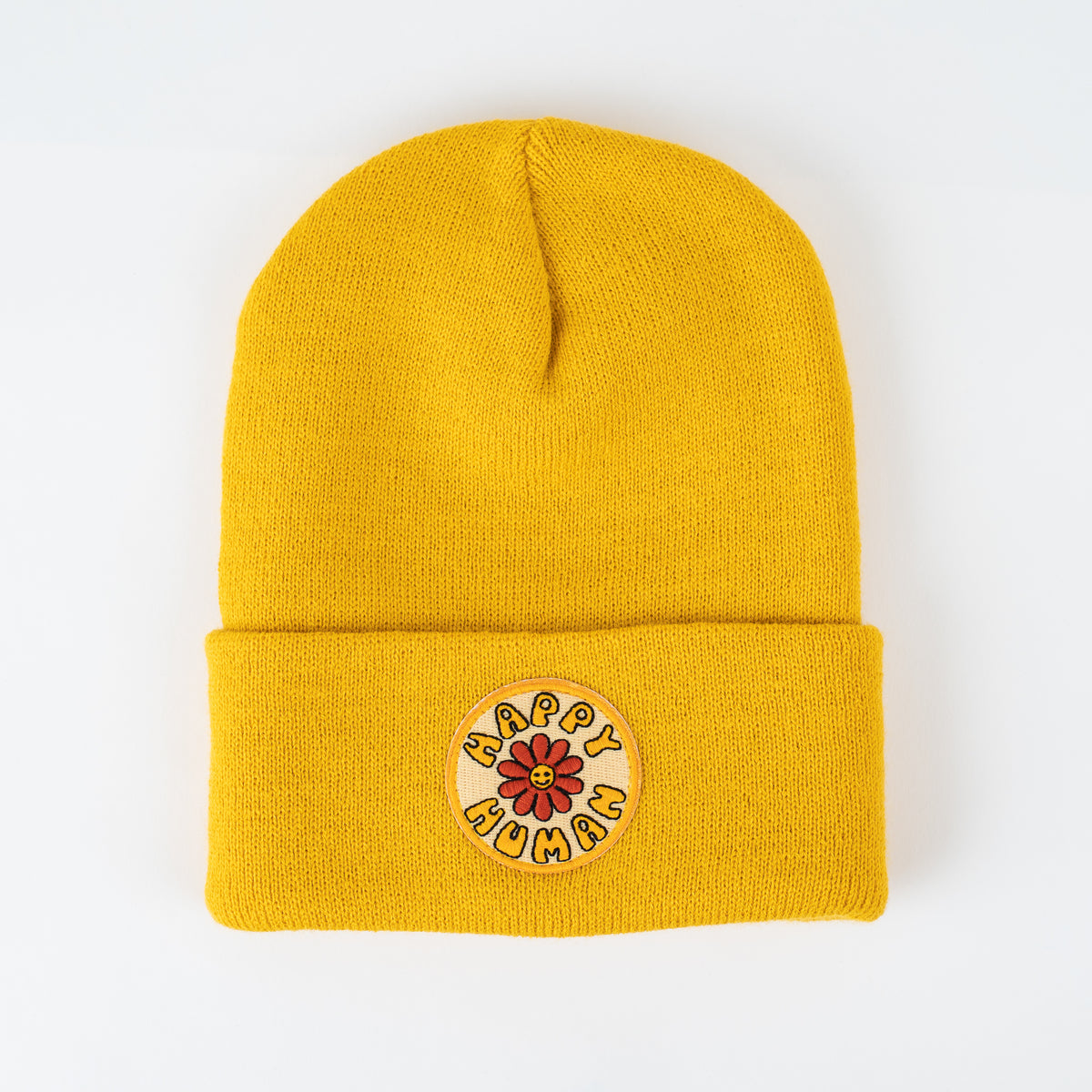 Introducing the "Happy Human | Sun - Adult Beanie." This delightful mustard yellow knit beanie features a folded brim and a round patch on the cuff, adorned with a red flower and the words "HAPPY HUMAN" in playful lettering. Displayed against a plain white background, it's an ideal choice for those who appreciate handcrafted style.