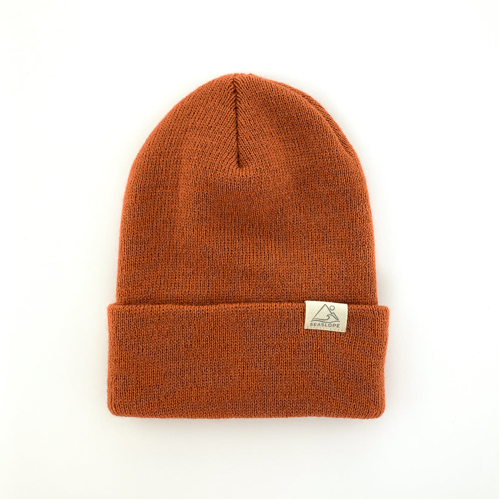 The CANYON INFANT/TODDLER BEANIE by seaslope1 is displayed on a simple white background. This knitted orange beanie, designed for toddlers, features a folded brim and includes a small rectangular patch.