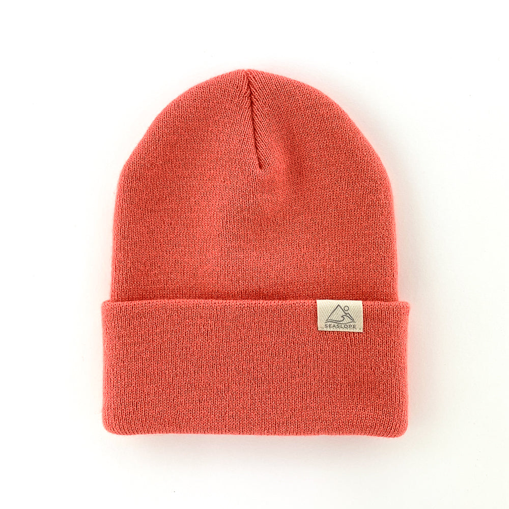 The Coral Infant/Toddler Beanie by Seaslope1 is a vibrant knit hat in coral, designed with a soft stretch fit. It features a folded brim and includes a small white rectangular patch with a triangle design. Made in the USA, this beanie is beautifully presented on a plain white background.