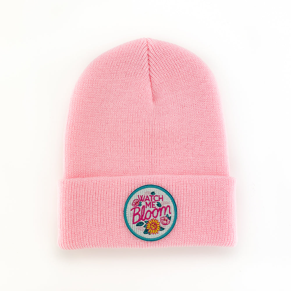 The WATCH ME BLOOM PEONY INFANT/TODDLER BEANIE features a folded brim, is made of 100% acrylic, and includes an embroidered circular patch with floral designs.