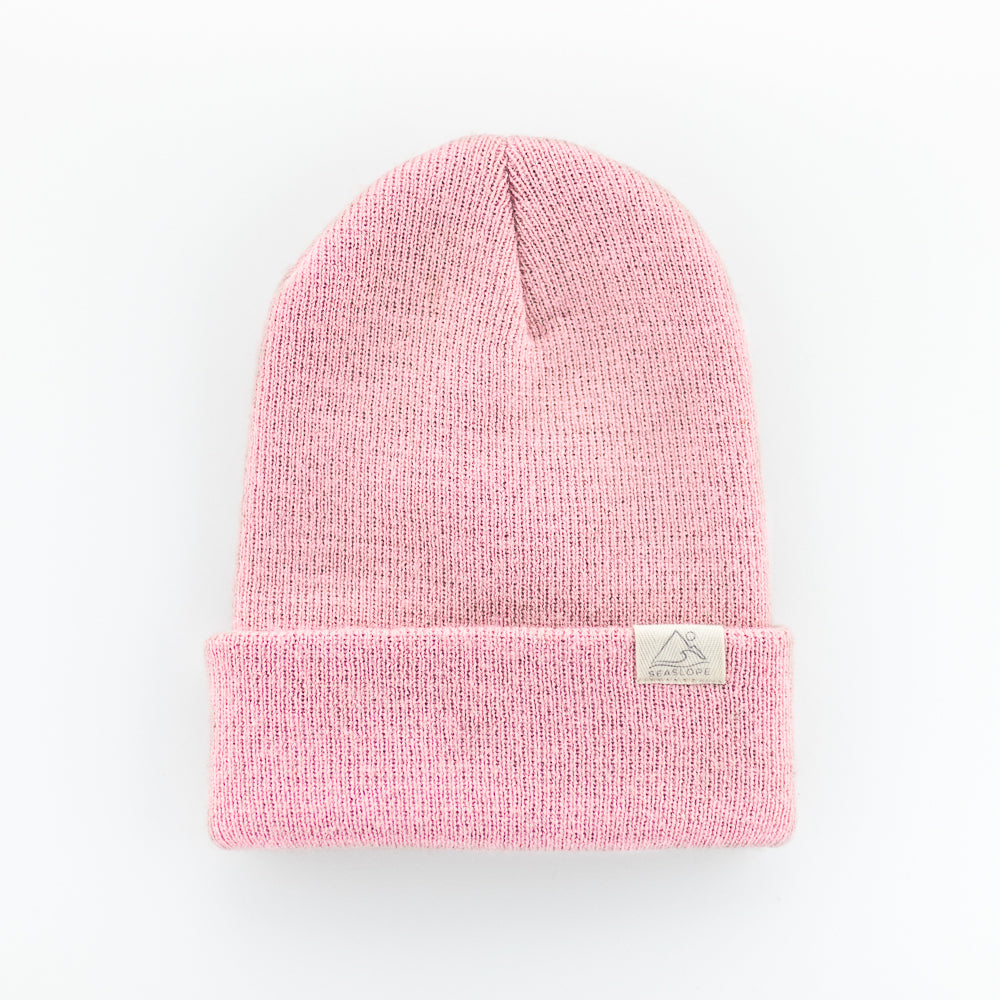 The PEONY INFANT/TODDLER BEANIE is a pink knit hat with a folded brim and a small rectangular label on the front. Made from 100% acrylic, it offers both style and comfort. This piece is proudly crafted in the USA and displayed on a plain white background for a clean look.