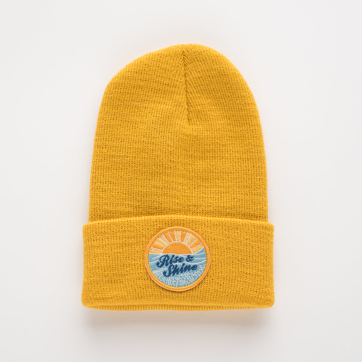 The Rise & Shine - Sun Kids Beanie is a mustard yellow acrylic beanie with a folded cuff. It features a unique round embroidered patch on the front, showcasing a sunrise design along with the words "Rise & Shine.