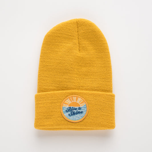 The Rise & Shine - Sun Kids Beanie is a mustard yellow acrylic beanie with a folded cuff. It features a unique round embroidered patch on the front, showcasing a sunrise design along with the words "Rise & Shine.