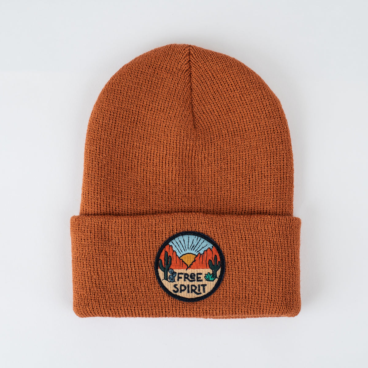 Introducing the "Free Spirit - Canyon Kids Beanie": This rust-colored beanie is crafted from 100% acrylic. It features an embroidered circular patch that reads "Free Spirit," depicting a desert landscape with cacti, orange mountains, and a blue sunburst against a light background.