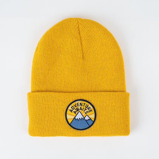 A cozy toddler beanie titled "Adventure Awaits - Sun Baby & Kid Beanie" comes in a mustard yellow shade. It features a folded brim and a circular patch on the front center with the phrase "Adventure Awaits" above illustrated mountains and a sun.