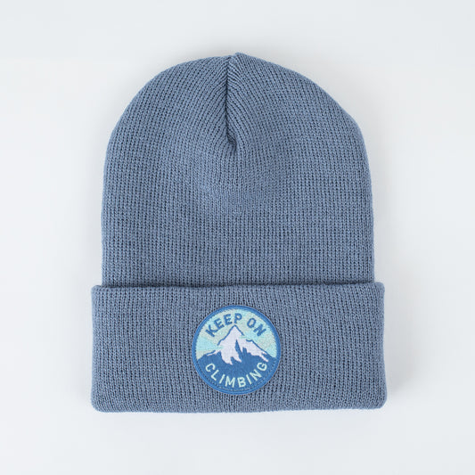 The Keep On Climbing - Pacific Kids Beanie is a blue knit beanie made in the USA from 100% acrylic. It has a folded brim and showcases a round patch with a mountain design, accompanied by the words "Keep On Climbing" embroidered around it on a plain light-colored background.