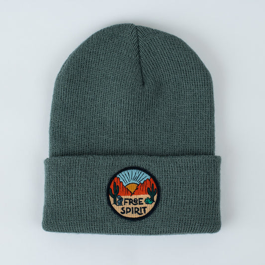 The infant/toddler beanie is a green acrylic knit with a folded brim. It features an embroidered patch depicting a desert scene with cacti, mountains, and a sunset, along with the words "Free Spirit" elegantly stitched below.