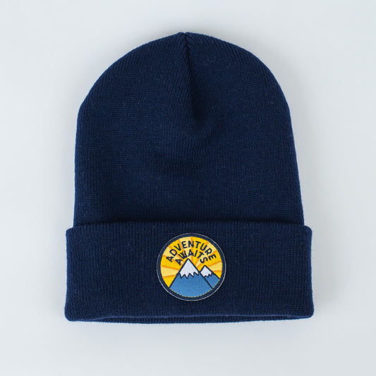 The beanie features a yellow embroidered patch with two blue mountains, highlighted by the phrase "ADVENTURE AWAITS" in bold black letters.