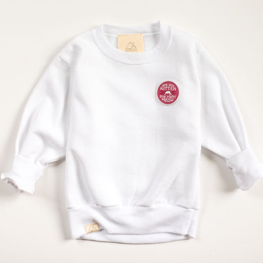 A cozy, long-sleeved white baby sweatshirt features a small embroidered red circular patch on the front with playful text. The soft fabric ensures comfort, and the label is neatly located at the neckline.