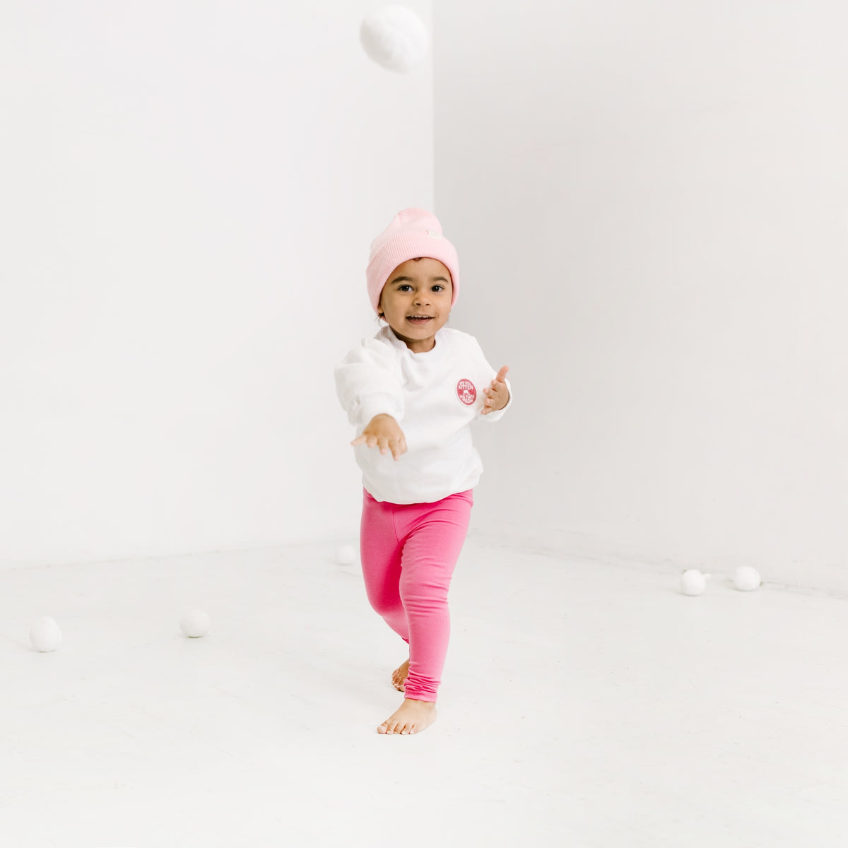 A toddler joyfully runs barefoot in a minimalist white room, wearing a cozy cotton sweatshirt with the phrase "Are You Kitten Me Right Meow," paired with pink pants and a matching pink beanie. The child seems to be having fun playing with white balls, and the sweater's embroidered patch adds a charming touch.