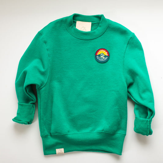 The GO EXPLORE SWEATSHIRT is a cozy green cotton crewneck with rolled sleeves. It features an embroidered patch depicting a mountain, rainbow, and the word "Outdoors," along with a small cream-colored tag on the bottom hem.