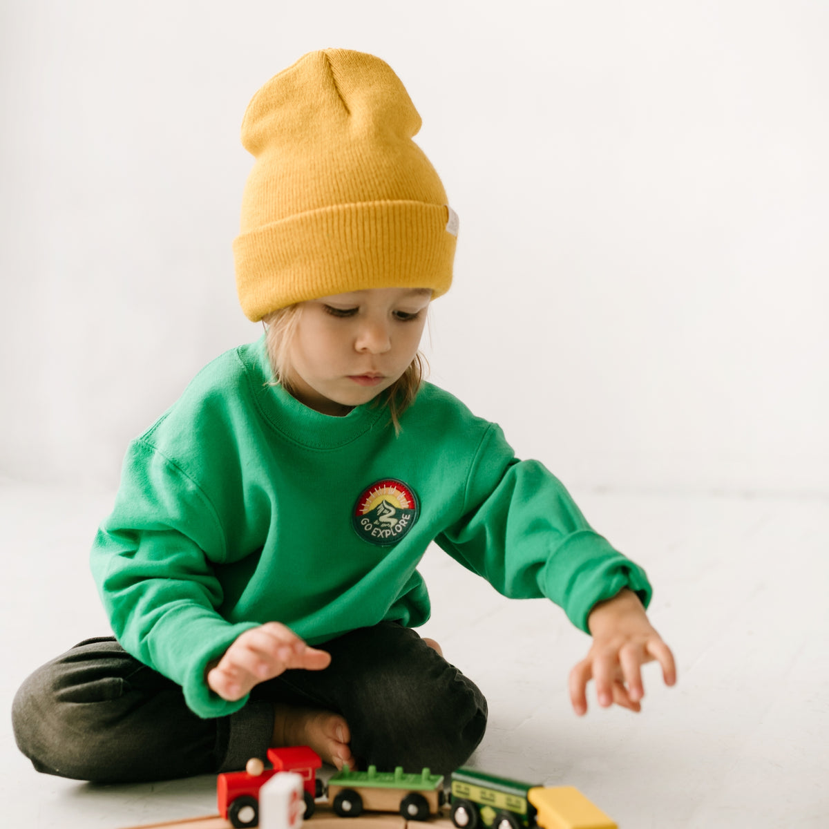 A child wears a cozy green cotton sweatshirt and a bright yellow beanie while playing with colorful wooden trains on the floor. The plain backdrop highlights this playful scene.