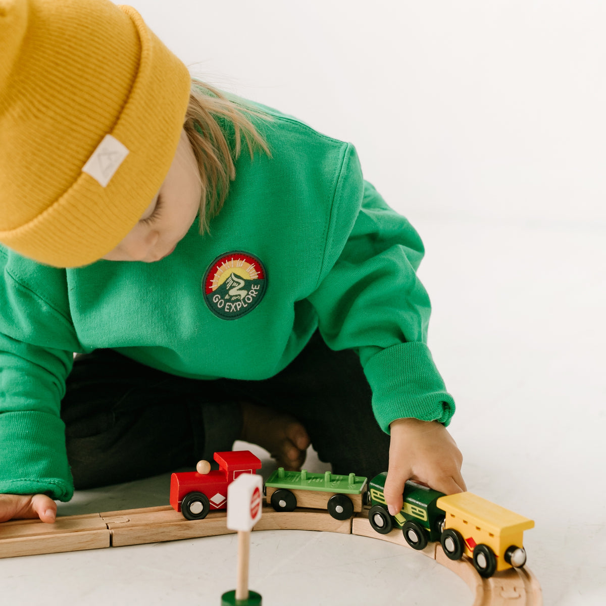 A child in a comfortable sweatshirt and a yellow beanie plays with a wooden toy train set on a light-colored floor. The bright red, green, and yellow train moves along curved tracks as the child focuses intently on arranging it.