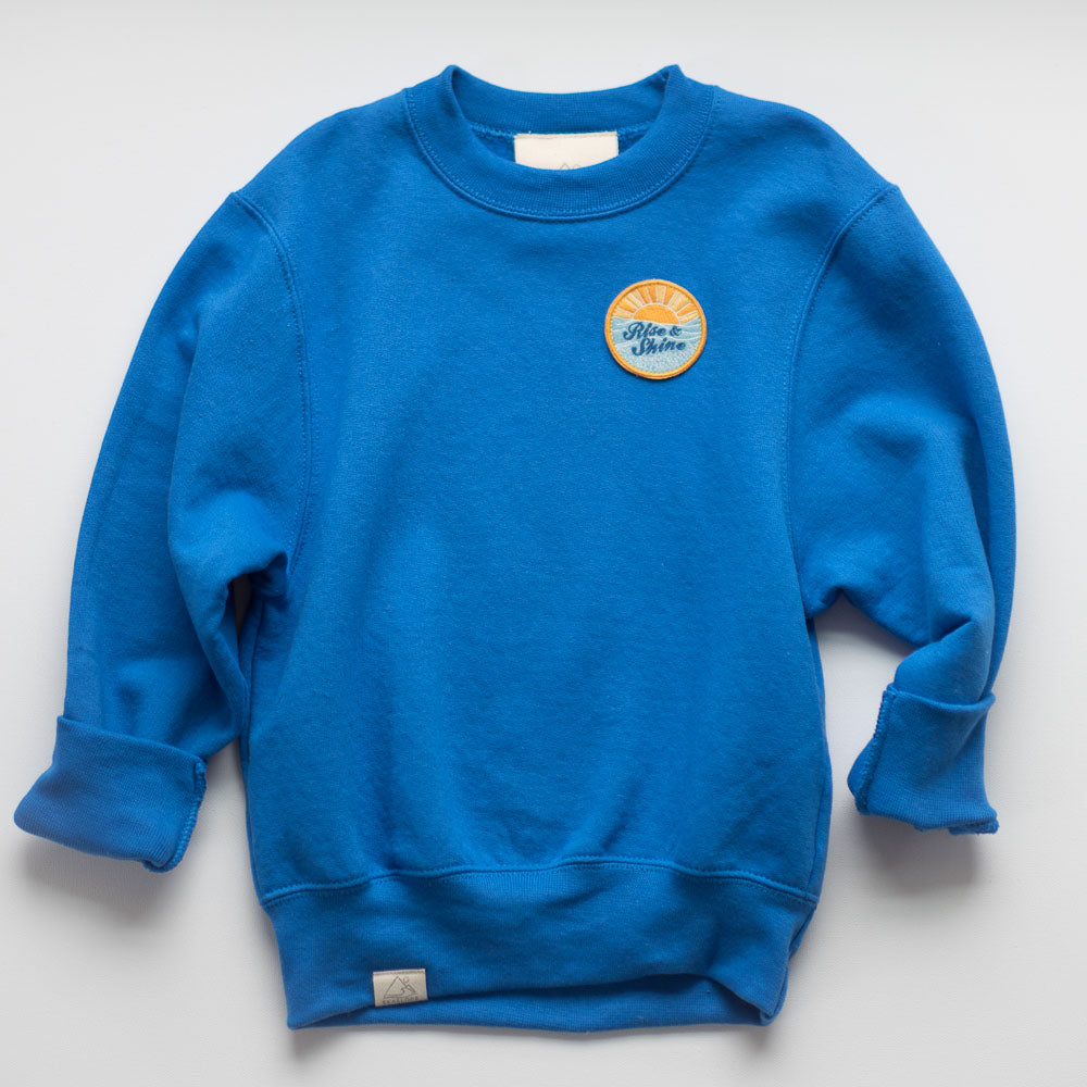 Introducing the RISE AND SHINE SWEATSHIRT: This ocean blue cotton pullover includes a round embroidered patch showcasing an orange sunburst design with the words "Rise & Shine." The sweatshirt features slightly rolled-up sleeves and a small fabric tag on the hem.