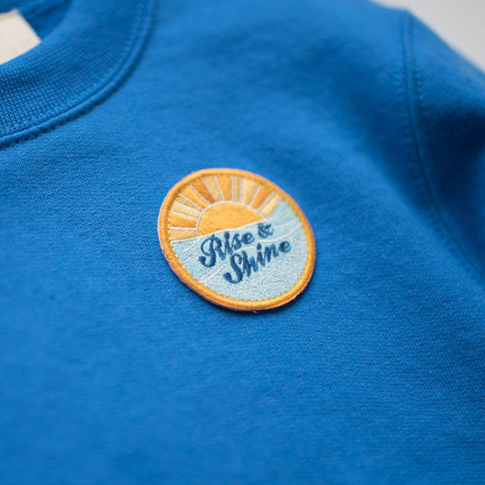 The RISE AND SHINE SWEATSHIRT by seaslope1 presents a stunning ocean blue color, accentuated by a distinctive round embroidered patch. This patch artistically illustrates a rising sun, accompanied by stylized script text saying "Rise & Shine." The embroidery skillfully combines vibrant shades of orange, yellow, and light blue.