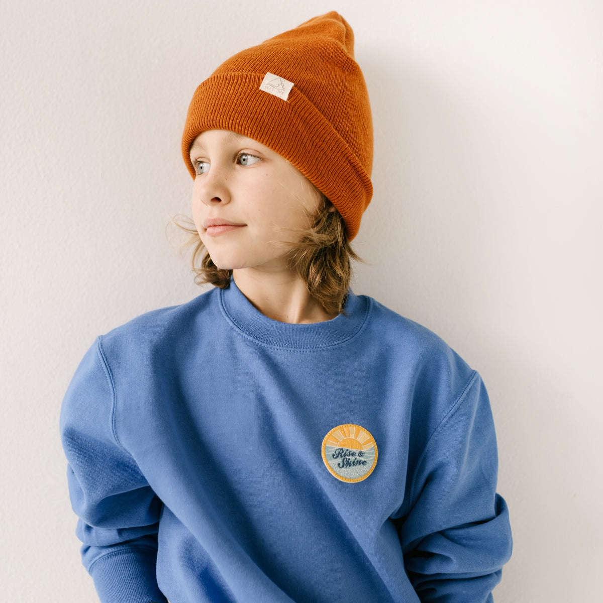 A young child is wearing an ocean blue cotton sweatshirt with an embroidered patch, looking to the side against a simple white wall.