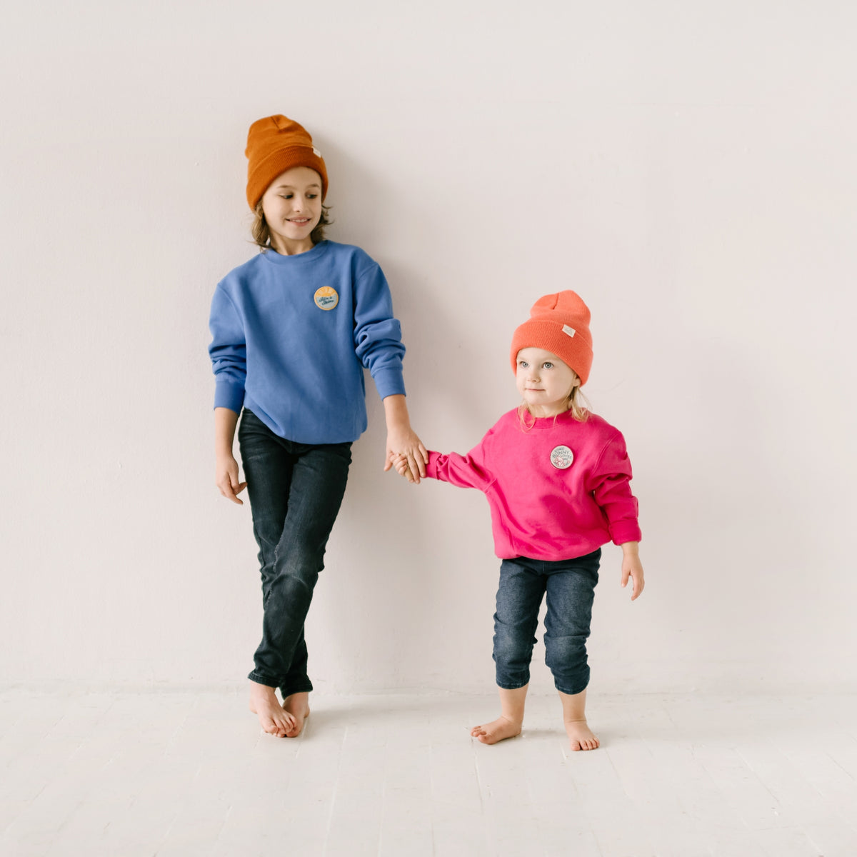 In front of a white wall, two children are holding hands. One is wearing an ocean blue sweatshirt paired with an orange beanie, while the other sports a pink sweater and matching beanie. Both are barefoot, displaying identical embroidered patches on their cozy apparel.