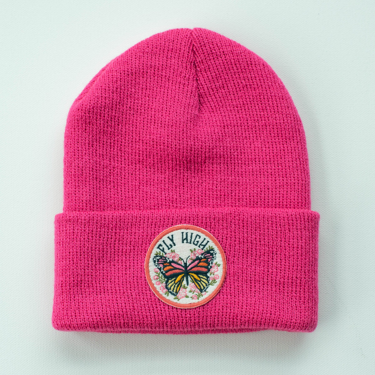 The fuchsia "Fly High Butterfly" kids beanie features a circular embroidered patch with a butterfly and floral design and the phrase "Fly High." This stylish accessory fits most sizes and is displayed on a light gray background.