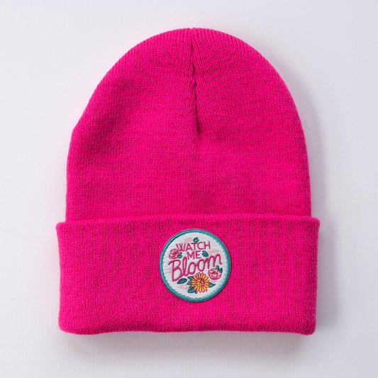 The vibrant fuchsia "Watch Me Bloom | Fuchsia - Adult Beanie" is a handmade knit accessory featuring a small, circular embroidered patch on the folded cuff. The patch displays the phrase "Watch Me Bloom" with colorful floral designs, creating a striking contrast against a simple, light background.