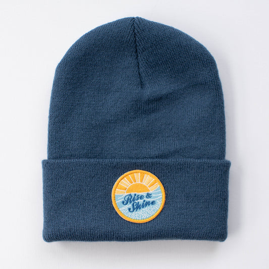 The Rise & Shine beanie is a blue knit hat with a folded brim. It features a circular patch depicting a sunrise over the water, with "Rise & Shine" written in blue script. This insulated, handmade accessory is ideal for keeping warm while adding inspiration to your morning style.