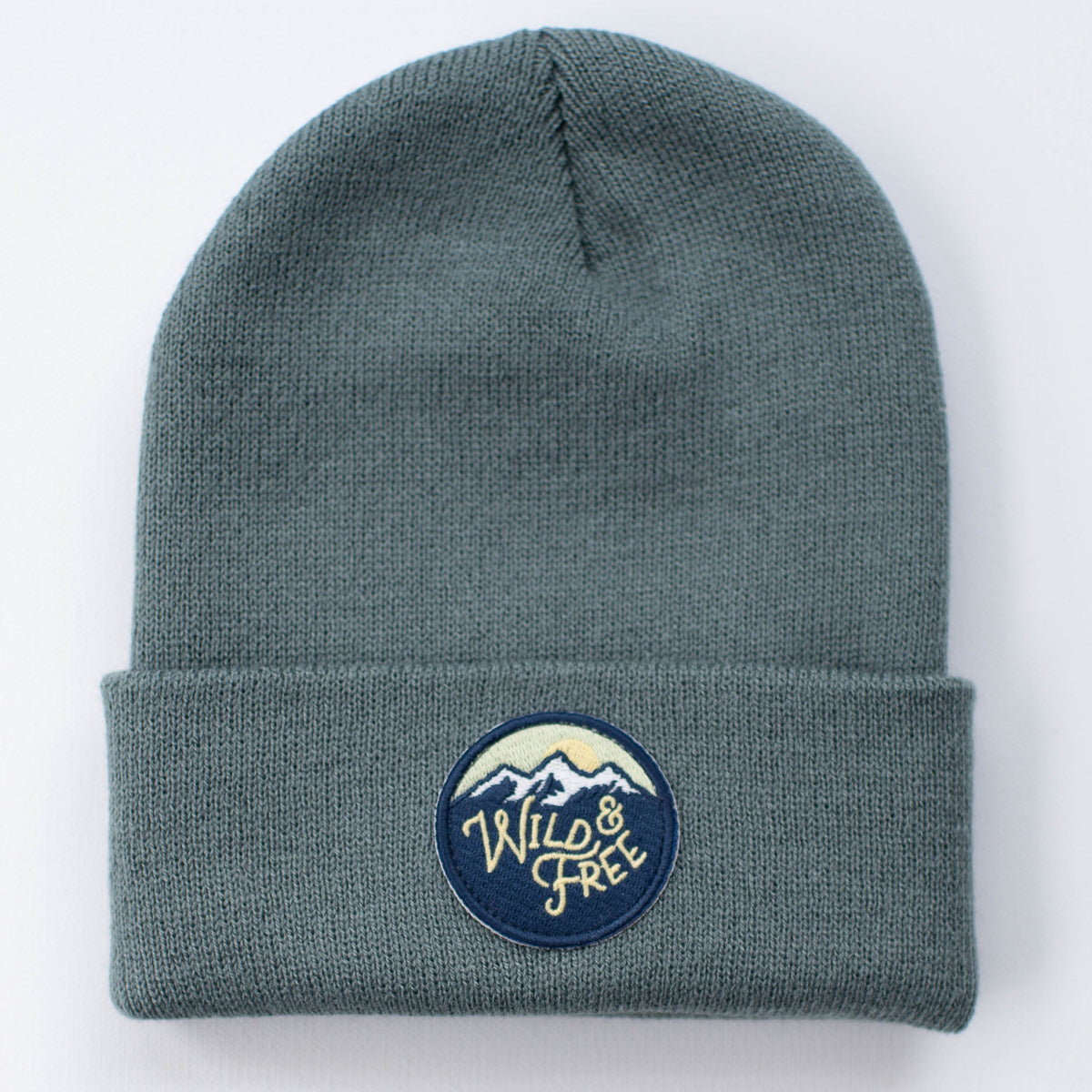 Introducing the WILD AND FREE SPRUCE YOUTH/ADULT BEANIE: a gray knit beanie with a folded brim, featuring a circular patch. This patch showcases an image of mountains accompanied by the words "Wild & Free" in stylized lettering.