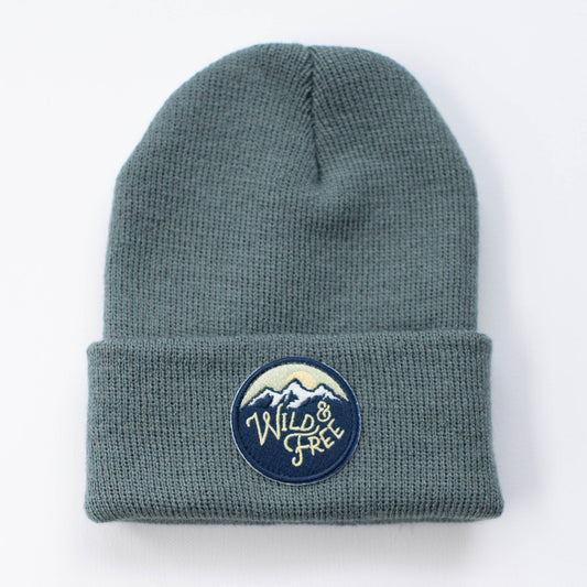 The Wild & Free - Spruce Kids Beanie from seaslope1 boasts a charming circular patch featuring a mountain design along with the text "Wild & Free" on its cuff, set against a crisp white background.