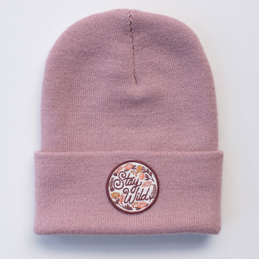 The STAY WILD ROSE YOUTH/ADULT BEANIE is a handcrafted pink hat known for its folded rim and detailed circular patch. The patch features the phrase "Stay Wild" and is adorned with floral and leaf motifs, showcasing beautiful embroidery in shades of white, pink, and orange.