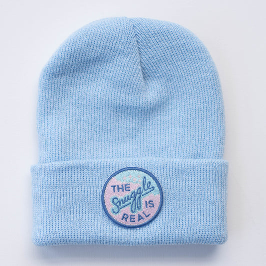 The SNUGGLE IS REAL Infant/Toddler Beanie by Seaslope1 is a light blue knit beanie featuring a folded brim. It's crafted from 100% acrylic material, with an embroidered patch displaying a pastel blue and pink circular design.