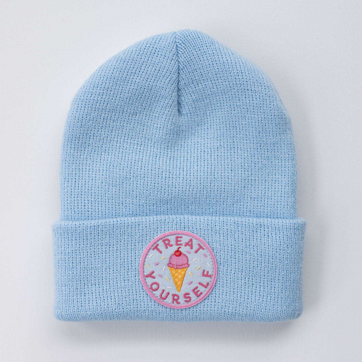 The TREAT YOURSELF SKY INFANT/TODDLER BEANIE by seaslope1 is crafted from 100% acrylic. It comes in a light blue shade and has a folded brim. The beanie is adorned with an embroidered circular patch that features an ice cream cone and the words "Treat Yourself" in pink.