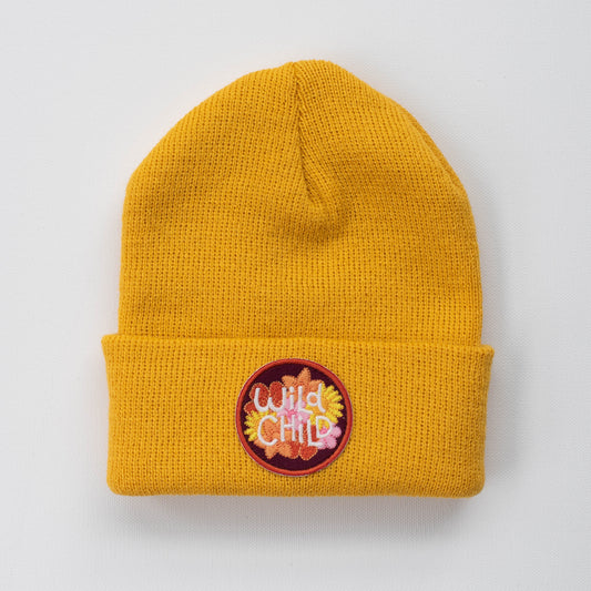 Introducing the Wild Child - Sun Kids Beanie: This cozy mustard yellow knit beanie is perfect for toddlers, featuring a stylish fold-over edge. The highlight is a circular patch with the words "Wild Child" embroidered in vibrant letters, framed by an orange and red border. Displayed against a plain white background, it perfectly showcases its charming design.